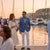 Man in a blue blazer and white pants standing next to a woman in a white dress by a marina at sunset.