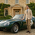 Man in a beige suit standing next to a vintage green sports car in front of a large house.